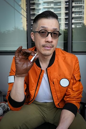 Photo of me dressed as Kim Kitsuragi from Disco Elysium. I'm a middle-aged Asian guy in an orange bomber jacket, olive pants, round glasses, and brown leather driving gloves. Sitting shot. I'm looking at the camera with a slight smile.