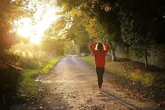 Woman wearing an orange sweatshirt and black pants walking along an autumn, tree-lined road, her hands on top of her head.