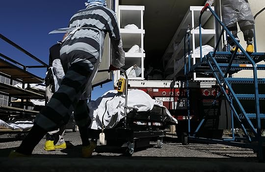 Low angle photo of a man in a black and white striped jail uniform pushing a cart with a dead body on it to a cargo truck stacked with other bodies.