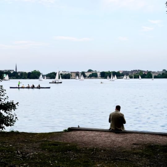Ein Mann sitzt am Ufer der Ufenalster, Sankt-Georg-Seite, er ist von hinten zu sehen. Vor ihm Ruder- und Segelboote, Sommreabend.