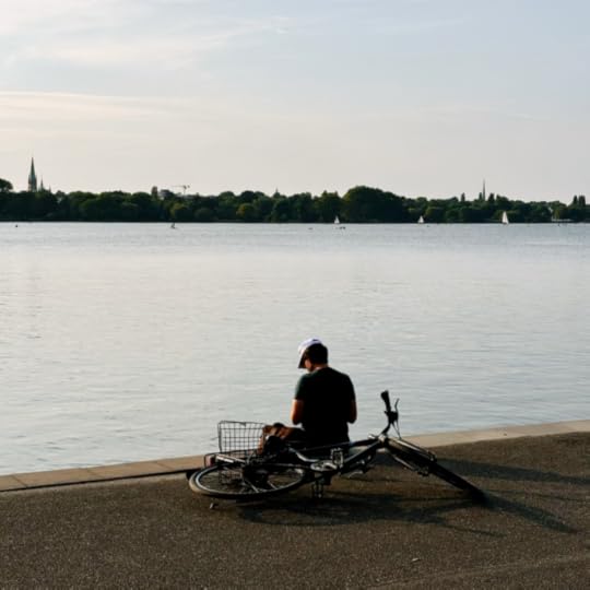Ein Mann sitzt am Ufer der Außenalster, Höhe Schwanenwik, er hat sein Fahrrad hinter sich abgelegt und liest etwas.