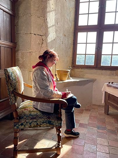 A person with reddish-brown hair tied back in a bun is seated on a wooden chair with floral upholstery, writing in a notebook. They are positioned by a large window, allowing soft, natural sunlight to flood the room. The sunlight casts shadows on the stone walls, enhancing the rustic and cozy atmosphere of the space. The person is wearing a white jacket and red hoodie, and they appear focused on their writing. The setting has an old-world charm with its worn stone walls, wooden furniture, and tiled floor. The scene conveys a peaceful, contemplative moment.
