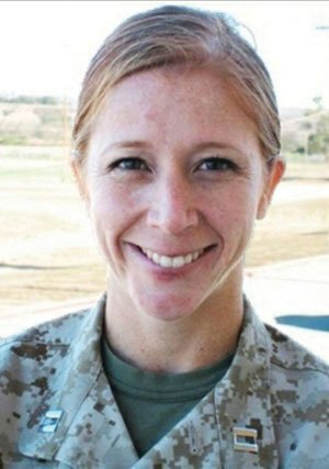 A person with light skin, blue eyes, and reddish-brown hair pulled back is smiling while wearing a military uniform with a camouflage pattern. The uniform has two rectangular insignia pins on the collar. The background shows an outdoor setting with indistinct structures and landscape