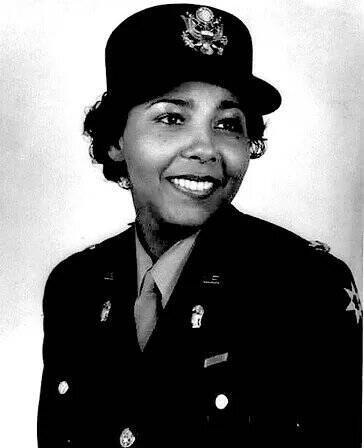 A black-and-white portrait of a smiling woman in a military uniform. She is wearing a peaked cap adorned with a badge and an emblem. Her buttoned-up jacket features insignias on the collar and has a row of buttons down the front. The background is plain and light-colored