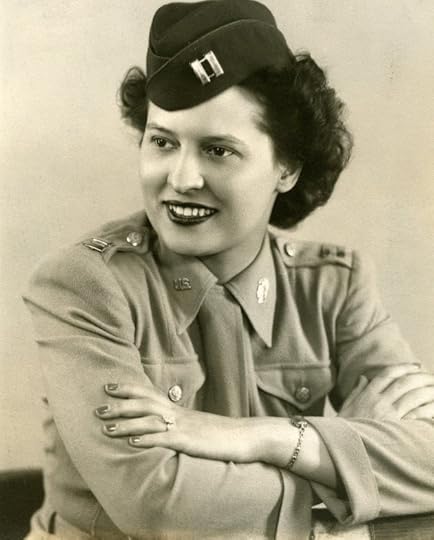 Black and white photo of a smiling woman in a military uniform. She is wearing a peaked cap with an insignia, a buttoned shirt with lapels, and a tie. Her arms are crossed along a surface, revealing a ring and bracelet on her left hand. Her curly hair is tucked under the cap.