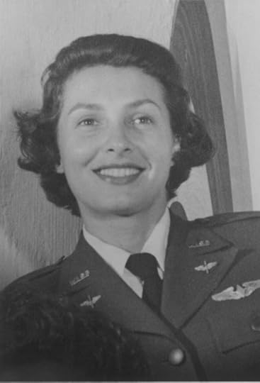 a grayscale photo shows a smiling woman with short, wavy hair. She wears a military uniform adorned with insignia and medals, including a winged badge above a nameplate. The woman appears confident and happy, standing against a lightly colored background.
