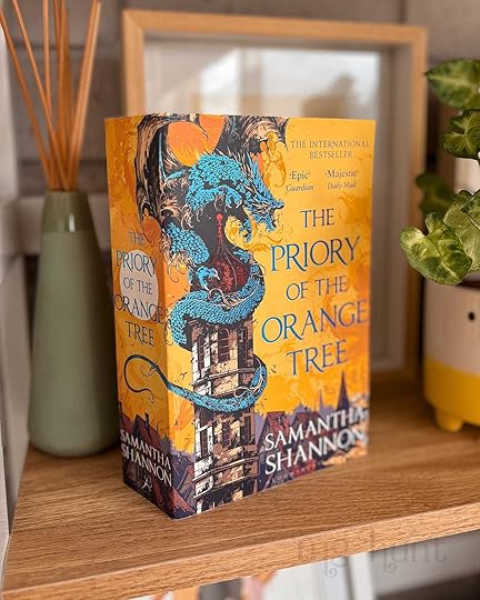 Book titled 'The Priory of the Orange Tree' by Samantha Shannon. The book is placed upright, on a slight angle, and it is sitting on a wooden shelf next to a pot plant and a reed diffuser.