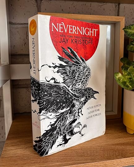 Book titled ’Nevernight’ by Jay Kristoff. The book is placed upright, on a slight angle, and it is sitting on a wooden shelf next to a pot plant and a reed diffuser.