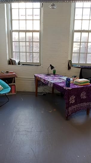 Artist studio with tall windows, table decorated with batiks and folders, bookscase with books, books in the window sills, reading chair