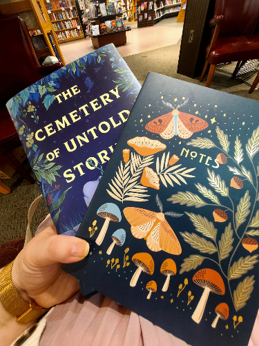Woman's hand holding a journal with mushrooms, leaves, and moths and a book with the title The Cemetery of Untold Stories