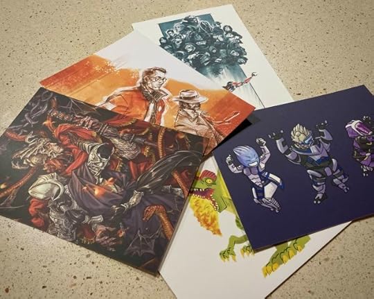 Photo of five postcards featuring my art laid out on the counter. We see Messmer and Rellana from Elden Ring, Kim and Harry from Disco Elysium, various characters from Mass Effect, and a dinosaur.