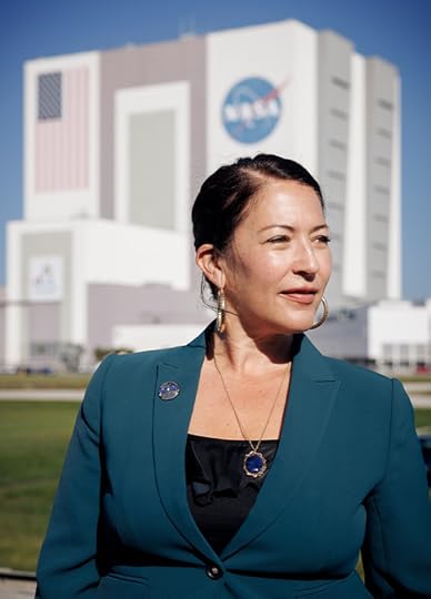 Color head and shoulders portrait of Ada Limon, facing to her left in bright sunshine with a NASA launch building in the distance.