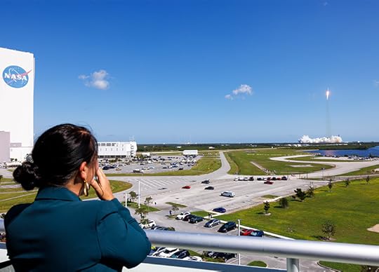 Wide shot of a woman leaning on a balcony, hands to her face, while in the distance a rocket trail can be seen high in the sky