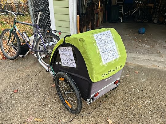 My bike, leaning against my open garage amid autumn leaves, with kid trailer attached, showing two flyers I have taped to the outside: 