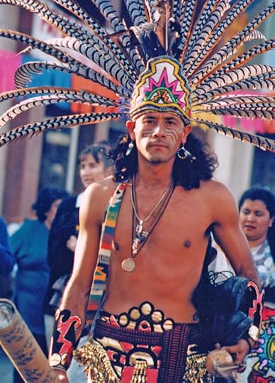 A thin, shirtless man wear an elaborate headress and colorful beads at a street festival faces the camera with a solemn expression