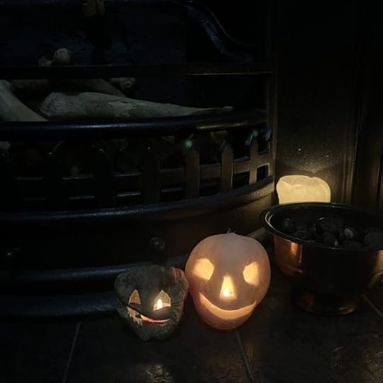 Photo shows a dark old cast iron fireplace full of bones, in front of it on the hearth is a lit candle, a brass bowl full of conkers and two turnip lanterns, one fresh and one shrivelled, both lit up.