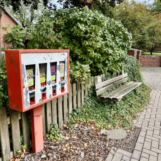 Alte und ramponiert aussehende Kaugummiautomaten neben einer zugewachsenen Sitzbank an einer Dorfstraße