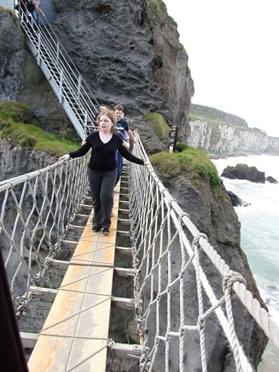 The image shows a narrow rope bridge with wooden planks, suspended over a rocky coastal area. A woman in a black sweater and dark pants is walking across the bridge, holding onto the rope handrails on either side. Behind her, a man is also crossing the bridge. The bridge is attached to a steep rocky cliff, and there is a metal staircase leading up the cliff on the left side of the image. In the background, you can see the ocean with waves crashing against the rocks and a distant view of more cliffs along the coastline. The sky is overcast, suggesting a cloudy day.