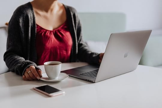 Woman wearing a red scoop necked blouse and a gray cardigan at a white table with a cup of tea, a cellphone, and a Macbook.