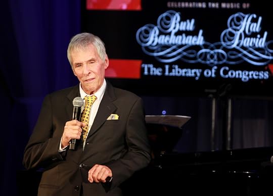 Bacharach, in his 80s, dressed in a black suit with a yellow tie, on stage. The red, blue and black screen behind him is illuminated with his name and that of his co-honoree, Hal David, written in white cursive script