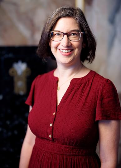 Half length color portrait of Aimee Hess. She's smiling, wearing glass and a red top, with shoulder-legth brown hair