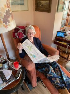 Elderly lady sitting in a coral coloured chair holding a paper. Her glasses are perched on the end of her nose and she’s smiling