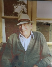 PIcture of an older man in a cardigan and shirt sitting in front of a window. He’s wearing a panama style hat and smiling.