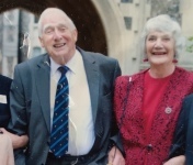 An elderly man and lady standing in front of a fling and sandstone archway. They are smartly dressed, her in a fuscia pink jacket and top, him in a suit with a striped dark blue and light blue tie. Their arms are linked and they are holding each other’s hands and smiling 