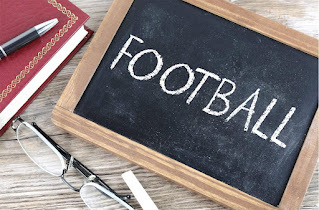 A small chalkboard on a desk. On the chalkboard, the word FOOTBALL is written. A pair of eyeglasses and a book with a red cover are also on the desk