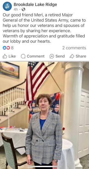 An image shows a group of older adults, both men and women, standing together on a stage in a hall. They are dressed in formal attire; the men are wearing suits with ties. An American flag is prominently displayed in the background next to a large blue curtain and a floral arrangement, suggesting this is a formal event or gathering.