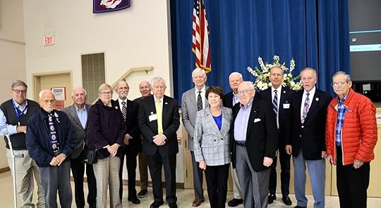 An image shows a group of older adults, both men and women, standing together on a stage in a hall. They are dressed in formal attire; the men are wearing suits with ties. An American flag is prominently displayed in the background next to a large blue curtain and a floral arrangement, suggesting this is a formal event or gathering.