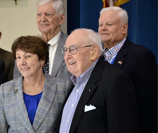 Five older adults, comprised of four men and one woman, stand together smiling against a formal backdrop. The lone woman wears a plaid blazer, while the men are dressed in suits; one man sports a pocket square. A blue backdrop and partial flag are visible in the background.