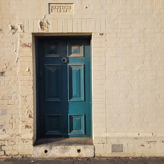 Photo of heavy teal door in a cream brick wall. The paint on the wall is flaking and the door is dusty. Above the door is the text 'Ola's home 1937'