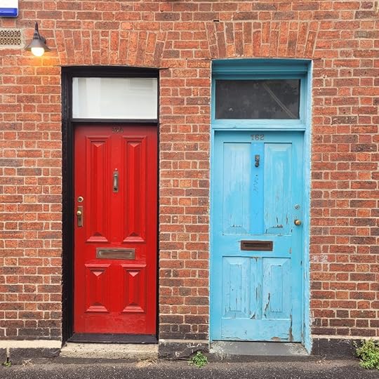 Red new door on left. Blue older, scuffed door on right. Both set in red brick wall.