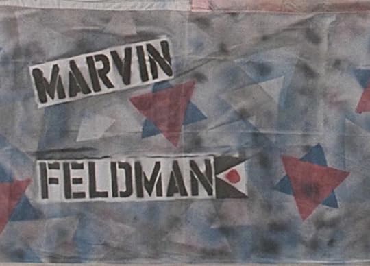 A dark gray section of a quilt, bearing the name Marvin Feldman in all capital letters, with geometric shspes in subdued reds and blues