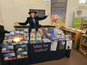 Picture of M T McGuire in a Pan of Hamgee style hat behind a stall selling her books in a St Edmundsbury Cathedral