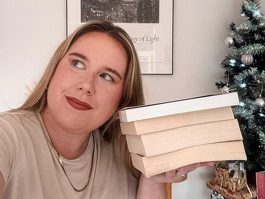 Author MG Hunt holding a stack of books with the pages facing the viewer. There is a white wall and a christmas tree in the background.
