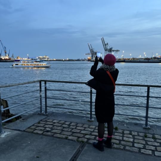 Die Herzdame, von hinten fotografiert, wie sie am Fischmarkt steht und über die Elbe mit dem Smartphone fotografiert. Später Nachmittag, es dunkelt schon, beleuchtete Fähren auf der Elbe, Hafenkräne im Hintergrund.