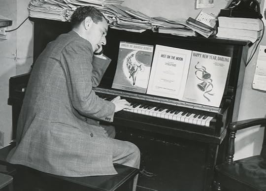 A middle-aged man dressed in a suit sits, turned sidways on a piano bench, chin in his left hand, right hand on the piano keys.