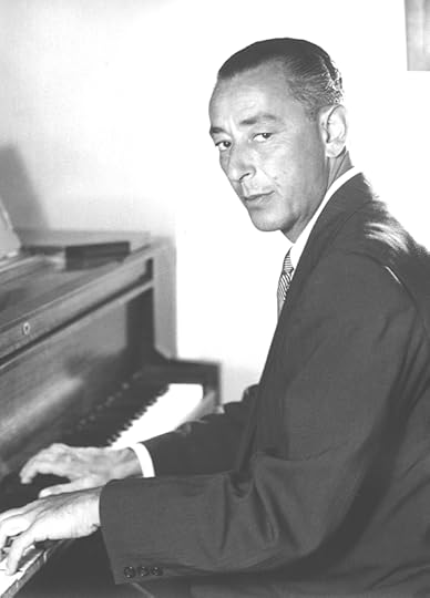 A middle-aged man in a suit sits at the piano with a serious expression, turned sideways to look at the camera.