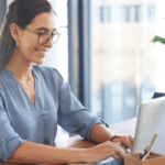 A woman wearing glasses is focused on her laptop, engaged in her work with a determined expression.