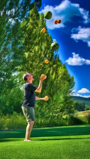a man juggling oranges and watermelons while standing on a lawn in front of trees