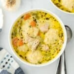 Overhead view of two bowls of chicken and dumpling soup with carrots, celery, and dumplings, accompanied by fresh herbs and garlic as garnish