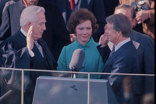 Medium close-up of Carter, right hand raised, taking the oath of office on a crowded platform.