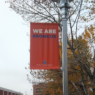 Image of an orange flag hanging from a silver pole outdoors. The flag reads: WE ARE BRONCOS