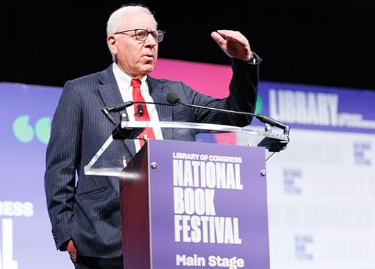 Half portrait of David Rubenstein, in a dark suit, white shirt and red tie, speaking at a podium with a purple 