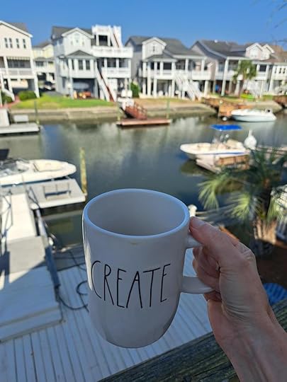 A white mug with the word create written in black print. In the background are palm trees, a dock and boats in the water. 