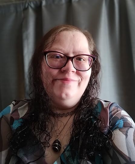 Picture of the author standing against a grey fabric background. The author is a white woman with a warm smile and large purple framed glasses, and a dimpled chin. She has long, fine, curly brown hair with scrunched curls. She is wearing a brown and blue patterned shirt, and a necklace.