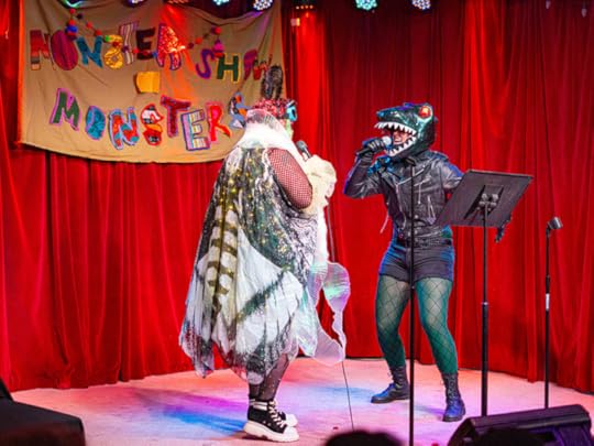 Two drag kings face off with microphones in hand, one dressed as Mothra and one dressed as Godzilla.