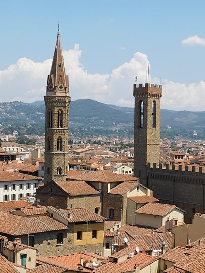 A photograph taken from my tower apartment across the roofs of Florence. Two golden stone towers stick up above all the red tiled roofs. On the left is a pointy one with crosses on it, part of the Badia monastery. On the right is a square one with battlements and big open windows, connected to a fortress with more battlements.